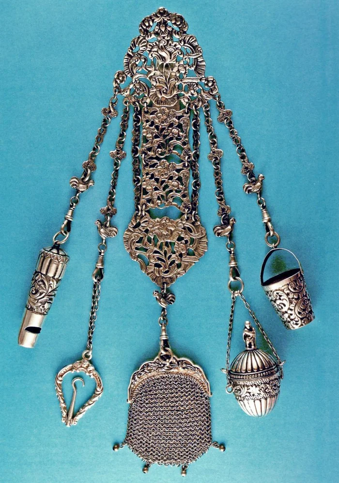 An ornate silver chatelaine laid flat, with mesh purse, bucket, and thimble case